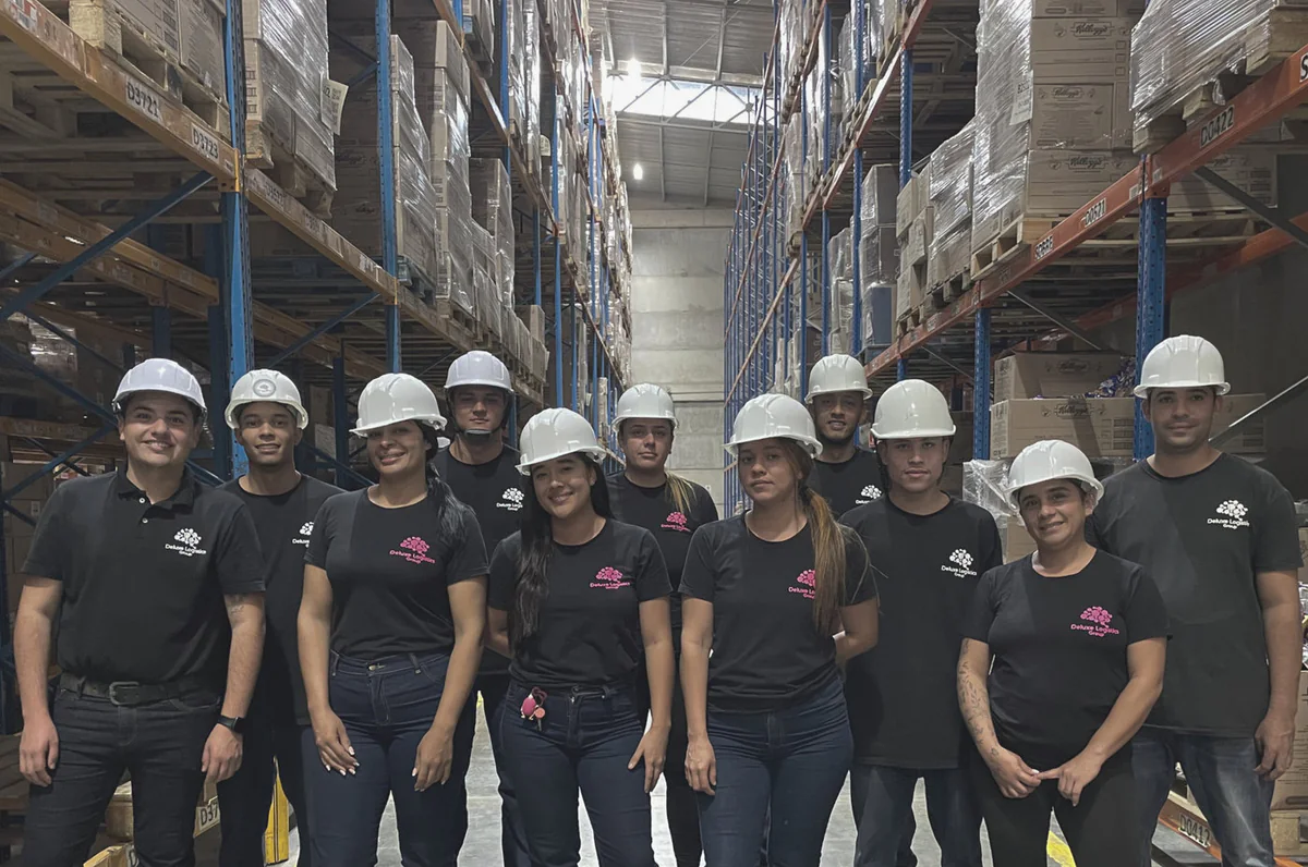 Colaboradores de Deluxe Logistics con casco blanco y camiseta negra de la marca, sonriendo en un pasillo de bodega con estanterías altas llenas de mercancía